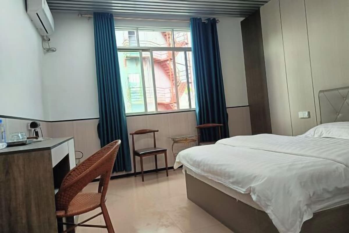 A hotel room with a bed, a desk, a chair, a window with curtains, and an air conditioning unit.
