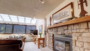 A cozy living room with a stone fireplace, a comfortable sofa, a television, and a large window offering a view of the outdoors. - Penthouse in Center Village, Extra Sleeping Area, Amazing views. (Copper Mountain)