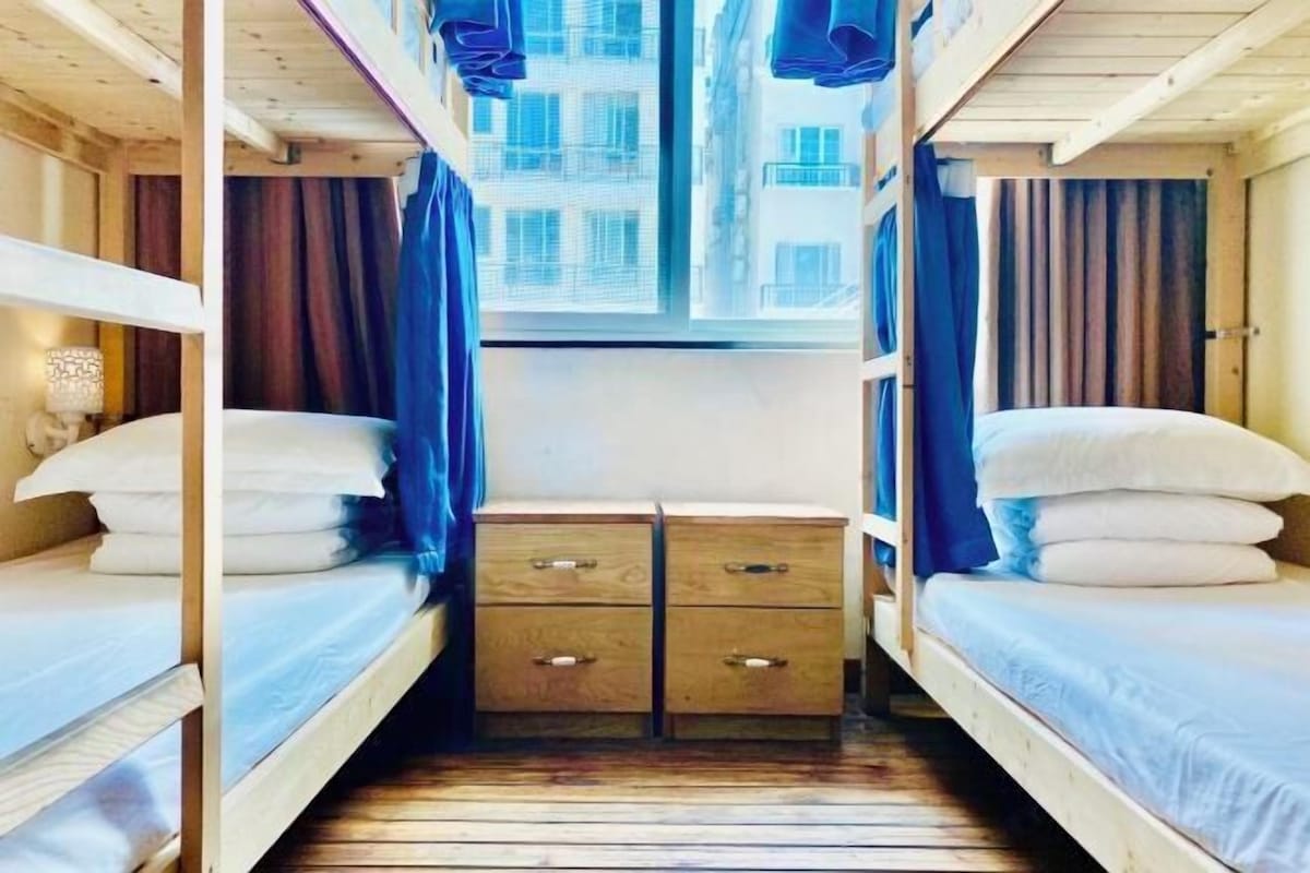 Een slaapkamer met een stapelbed, raam, gordijnen en een klein houten dressoir.