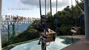 Eine Glasbrücke mit einer Schaukel, umgeben von viel Grün und Blick auf eine ferne Landschaft.