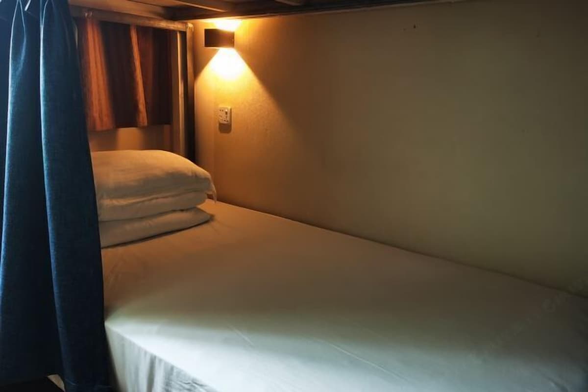 Een enkel bed met een witte matras en hoofdbord, een aan de muur bevestigde lamp en een gordijn dat gedeeltelijk is dichtgeschoven.