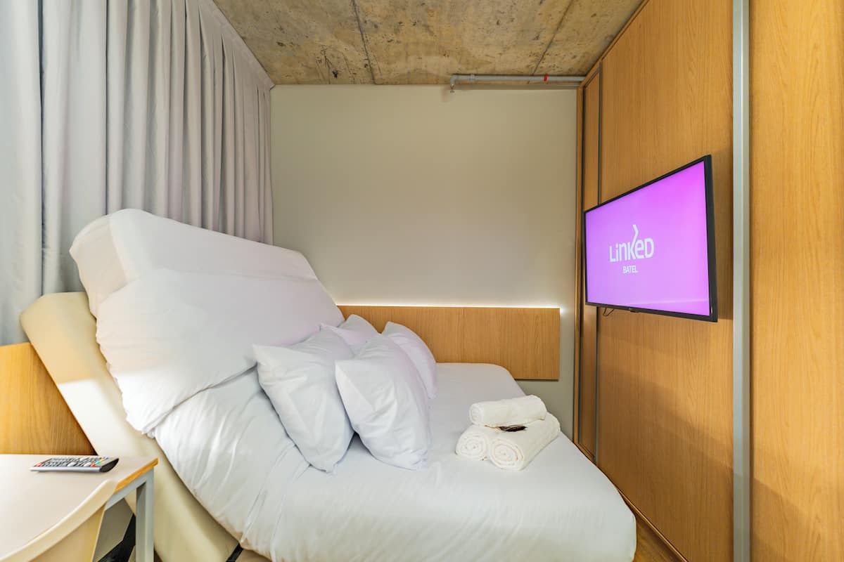 A hotel room with a bed, a TV mounted on the wall, a remote control on the bedside table, and a neatly made bed with rolled towels.