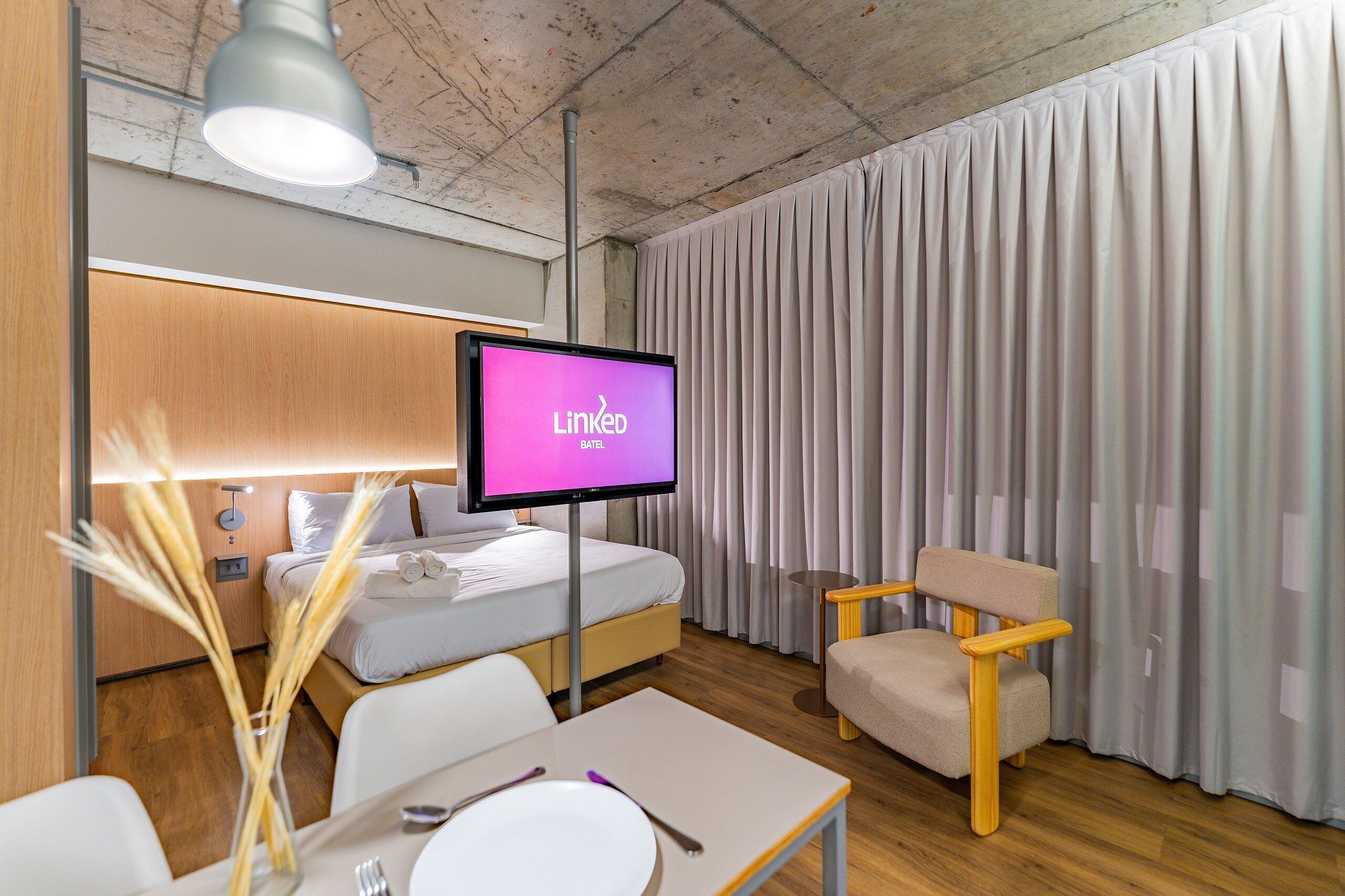 A hotel room with a bed, a TV displaying LinkedIn, a chair, a table with a plate and utensils, and a vase with reeds.