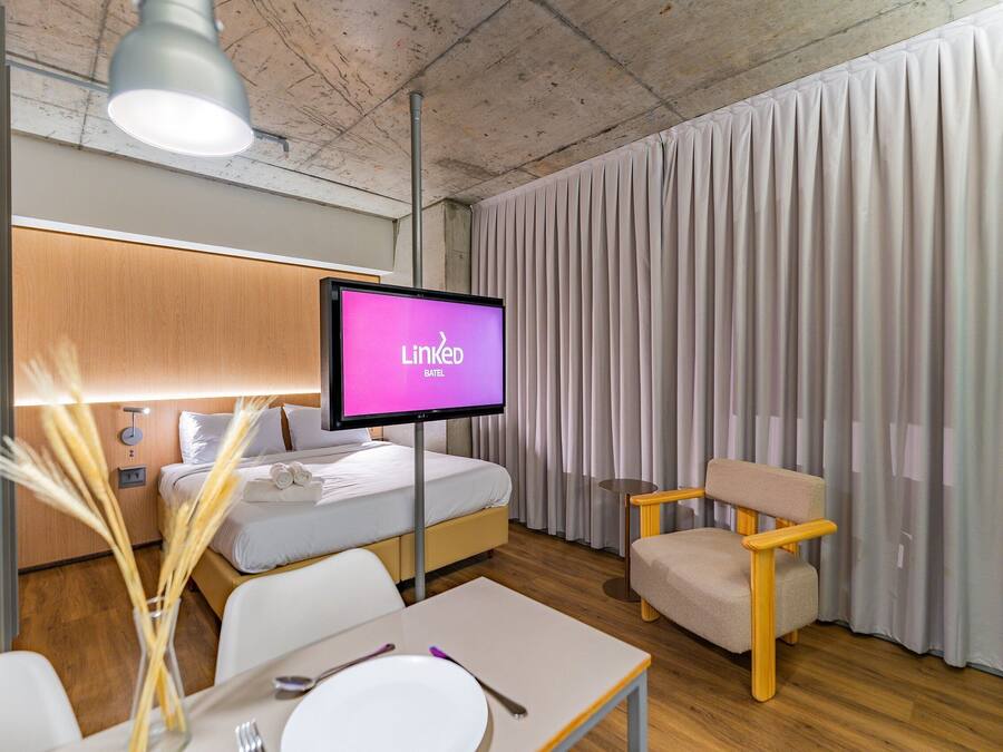 Habitación de hotel con cama, televisor mostrando LinkedIn, una silla, una mesa con plato y cubiertos, y un jarrón con cañas.