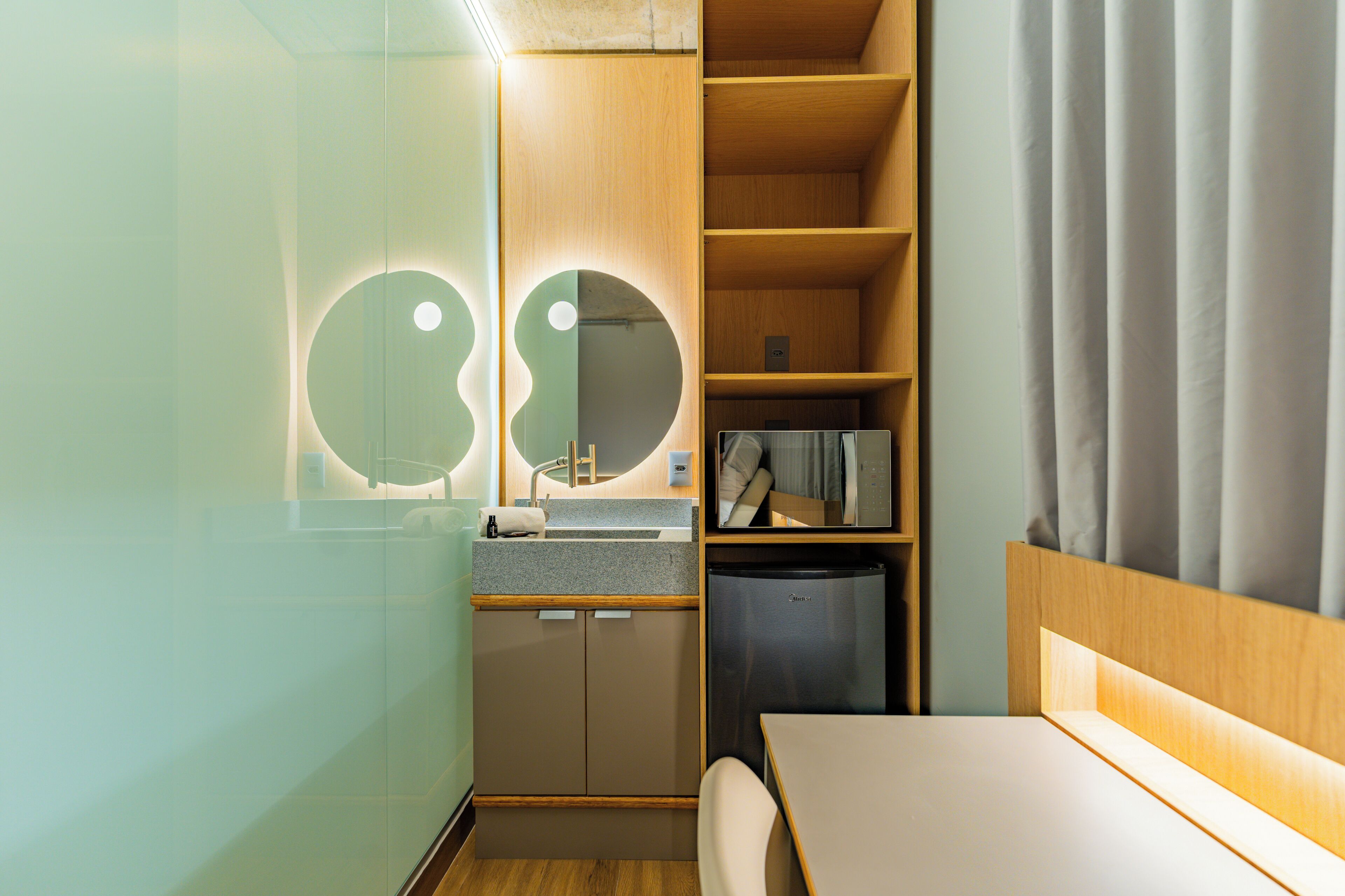 A compact kitchenette with a sink, a small refrigerator, and a mirror with circular light fixtures.