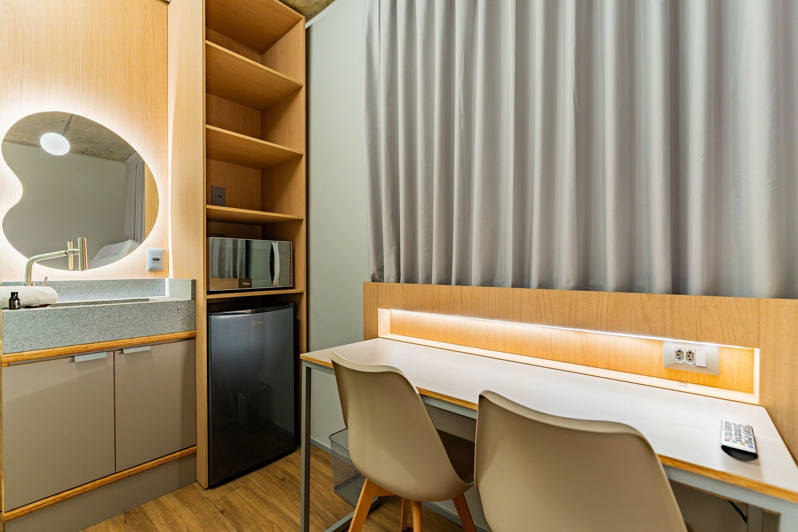 A compact hotel room with a kitchenette, a desk with two chairs, a built-in mirror, and a large window with curtains.