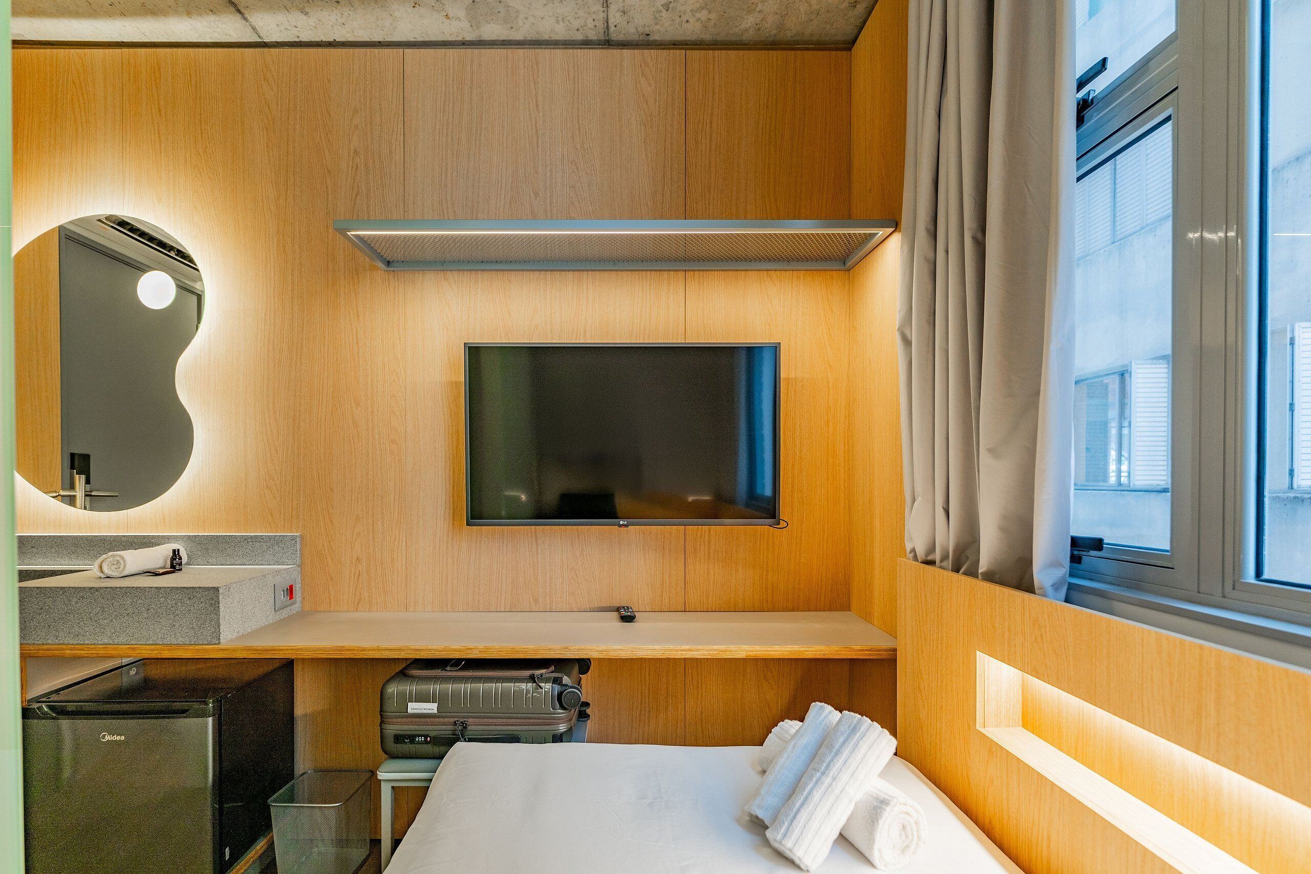 Habitació d'hotel amb llit, televisió penjada a la paret, petita cuineta i finestra amb cortines.