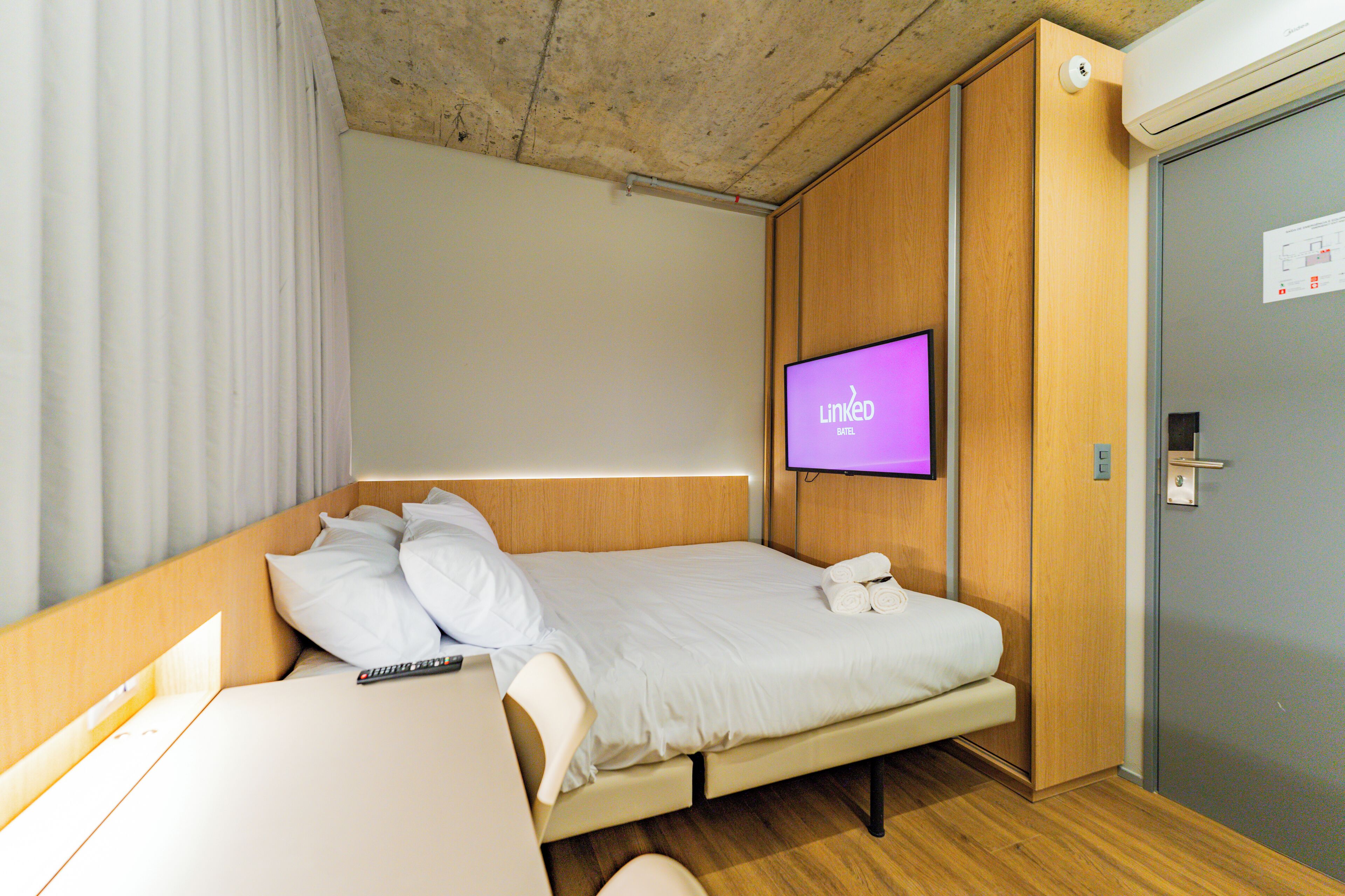 Quarto de hotel com cama, televisão fixada na parede, porta de madeira e janela com cortinas.