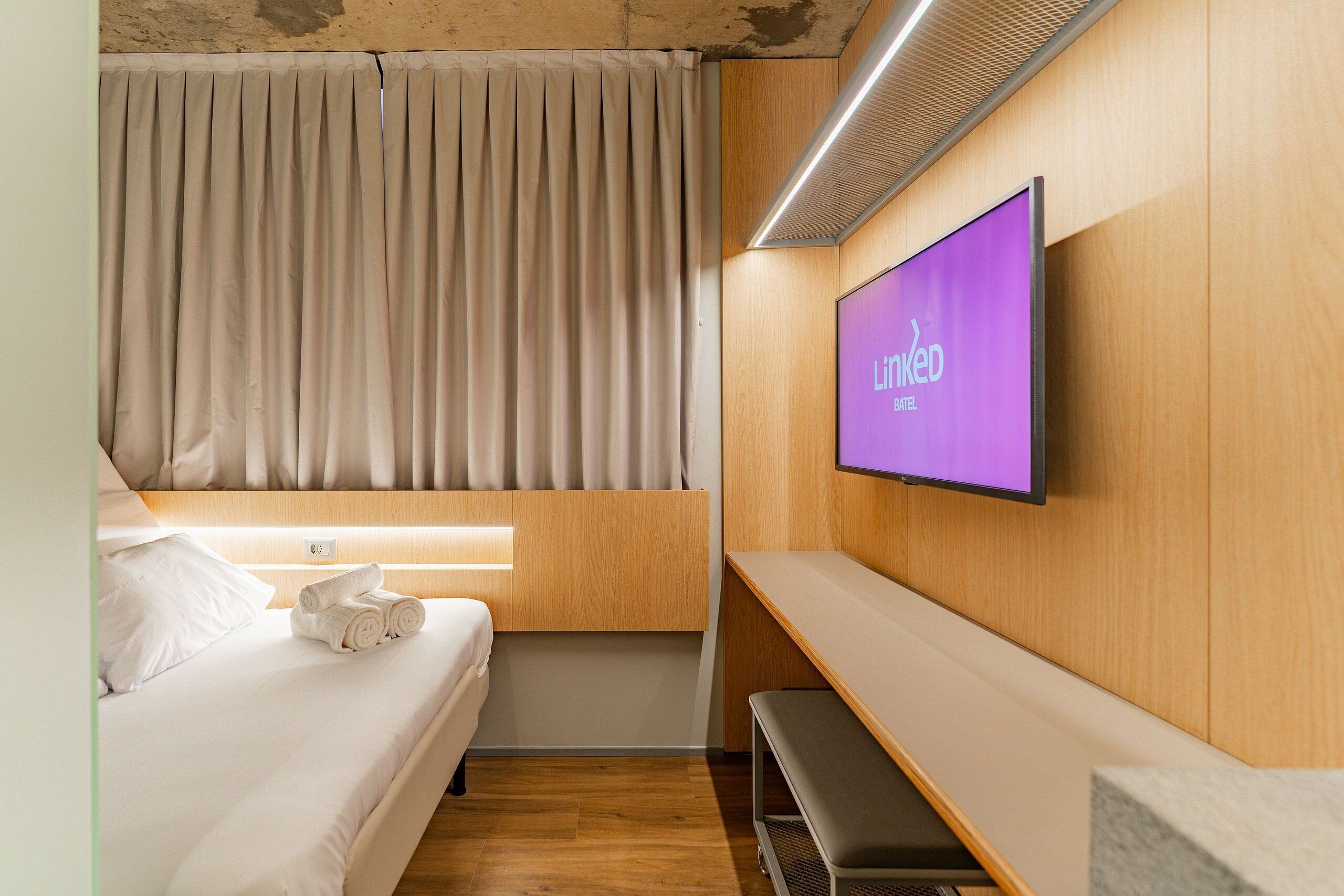 Quarto de hotel com cama, televisão fixada na parede, um banco e janela com cortinas.