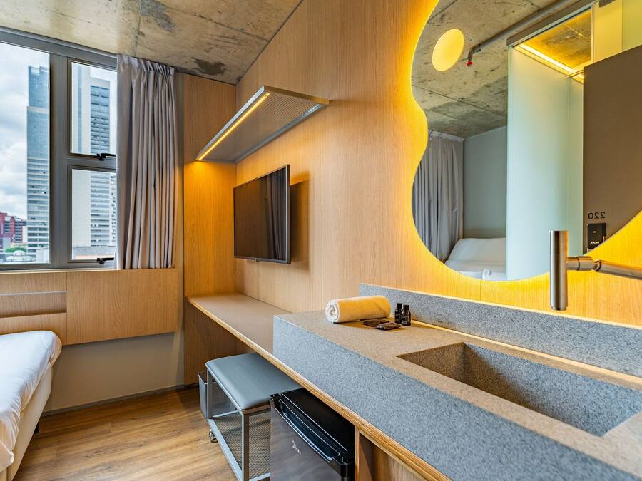 Una habitación de hotel moderna con un ventanal grande, un lavamanos integrado, una banca y un televisor de pantalla plana.