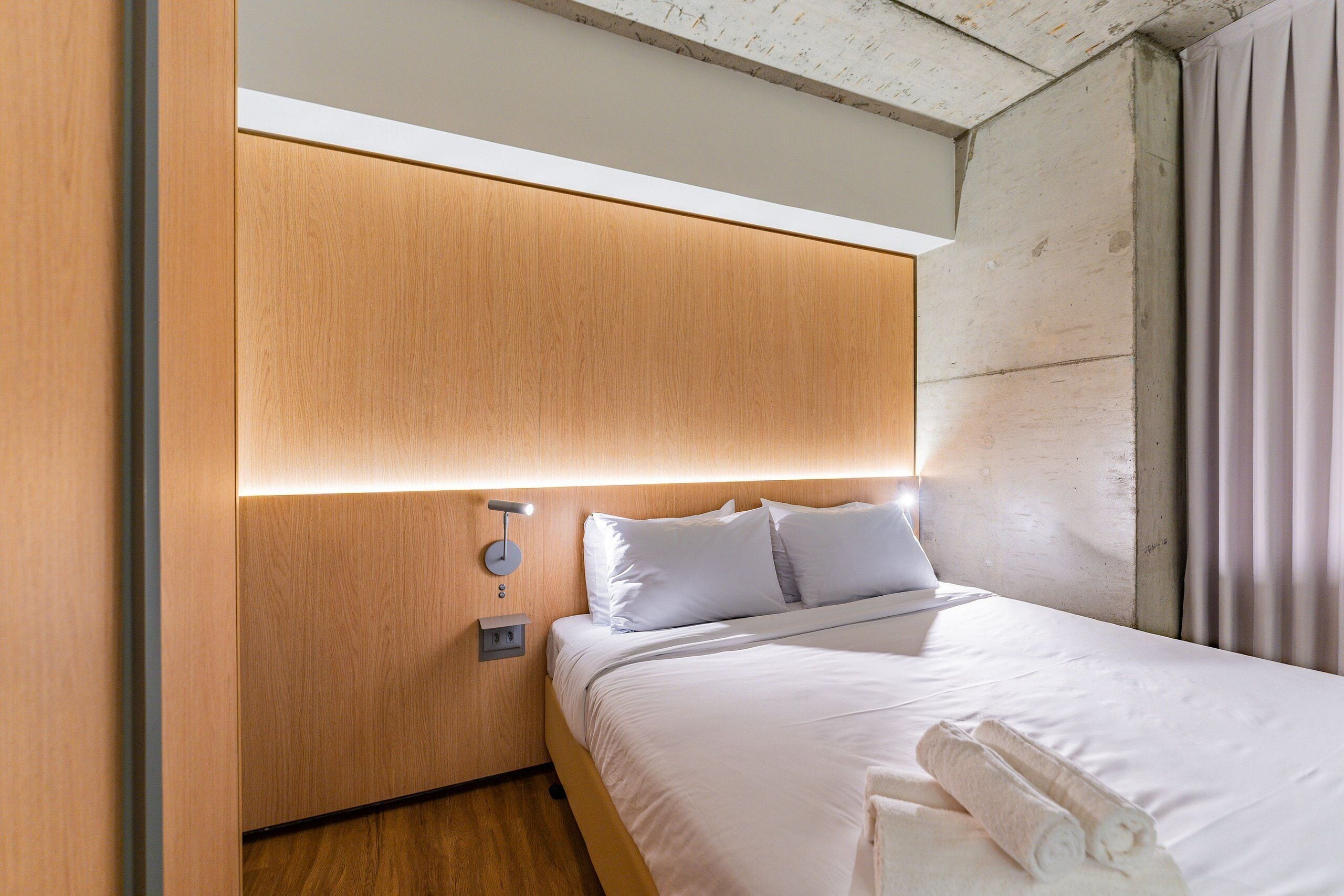 Quarto de hotel moderno com cabeceira de madeira, cama bem arrumada e luminária fixa na parede.