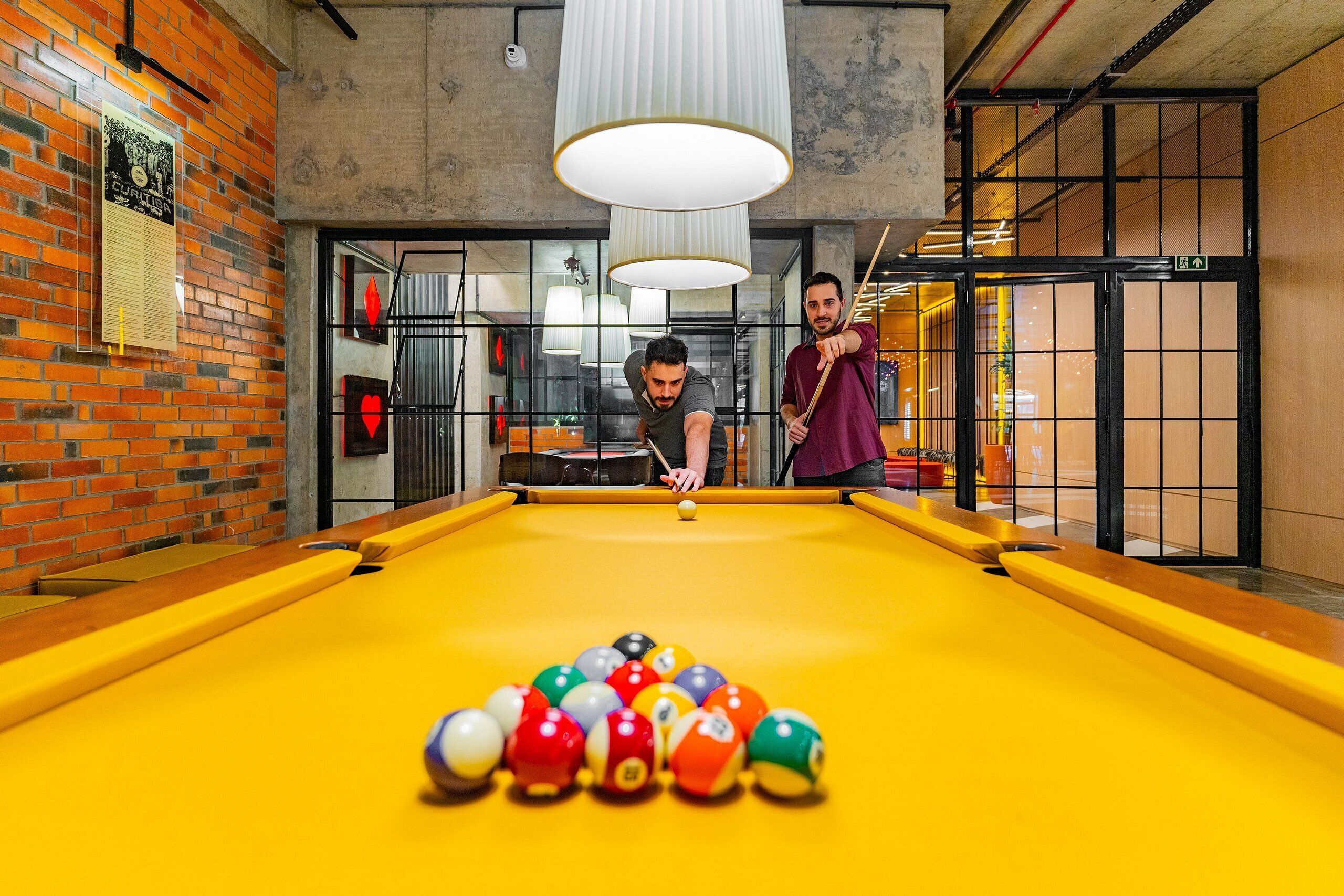 A pool table with balls and two people playing, a brick wall, and a large light fixture.