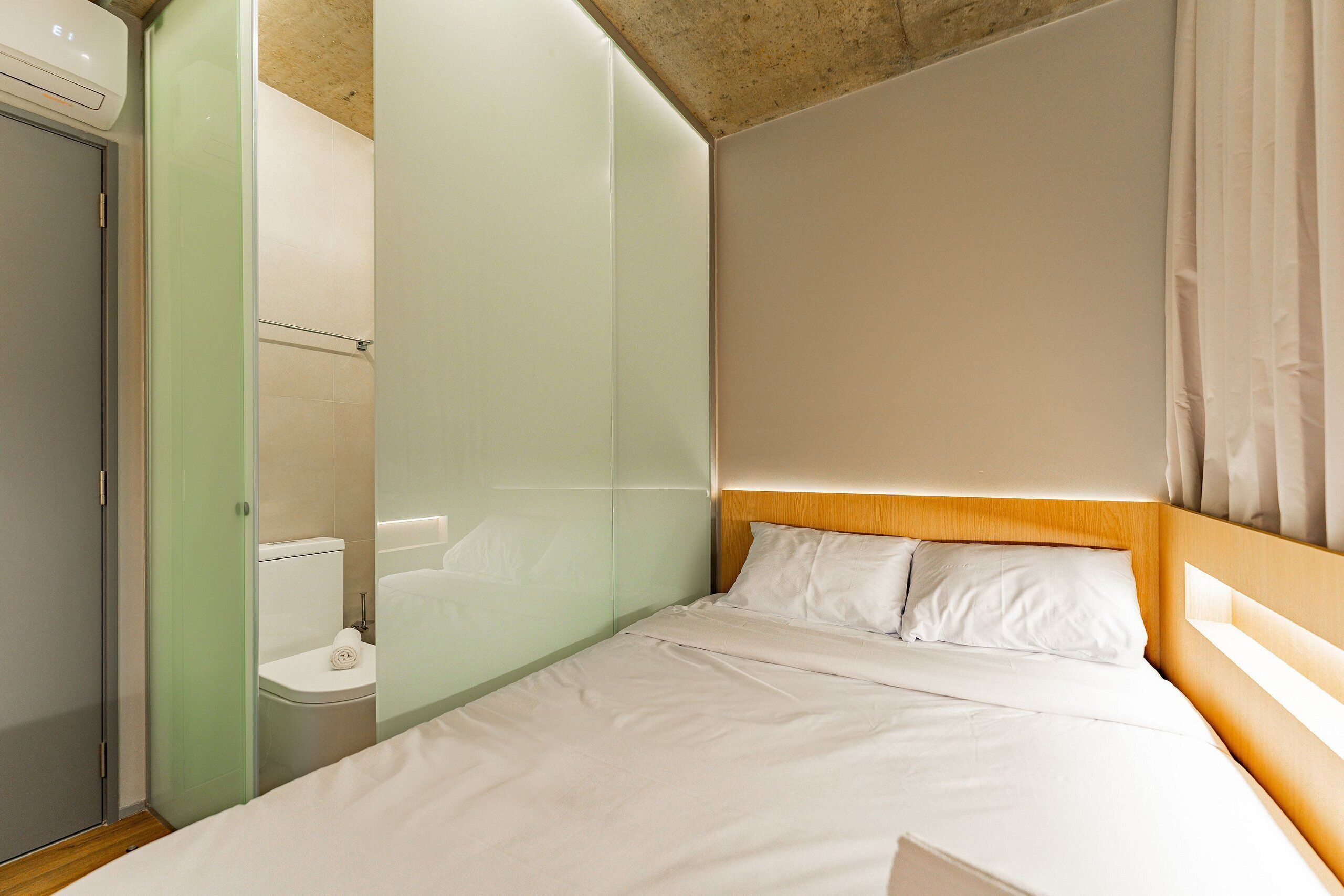A hotel room with a bed, a wooden headboard, a toilet, and a glass partition.