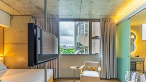 A hotel room with a bed, a TV, a chair, a small table, and a view of the city. - Linked Batel by Xtay (Curitiba)