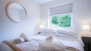 Una cama bien hecha con ropa de cama y toallas blancas, un espejo redondo en la pared, una ventana con vistas a la vegetación y dos lámparas de noche.