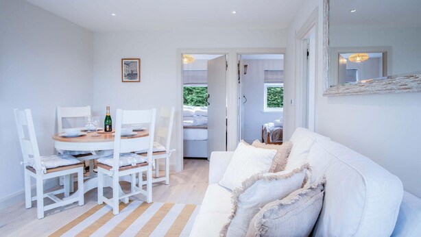 Una habitación moderna y luminosa con comedor que incluye una mesa redonda y sillas blancas, un sofá blanco con cojines decorativos y un espejo grande en la pared.