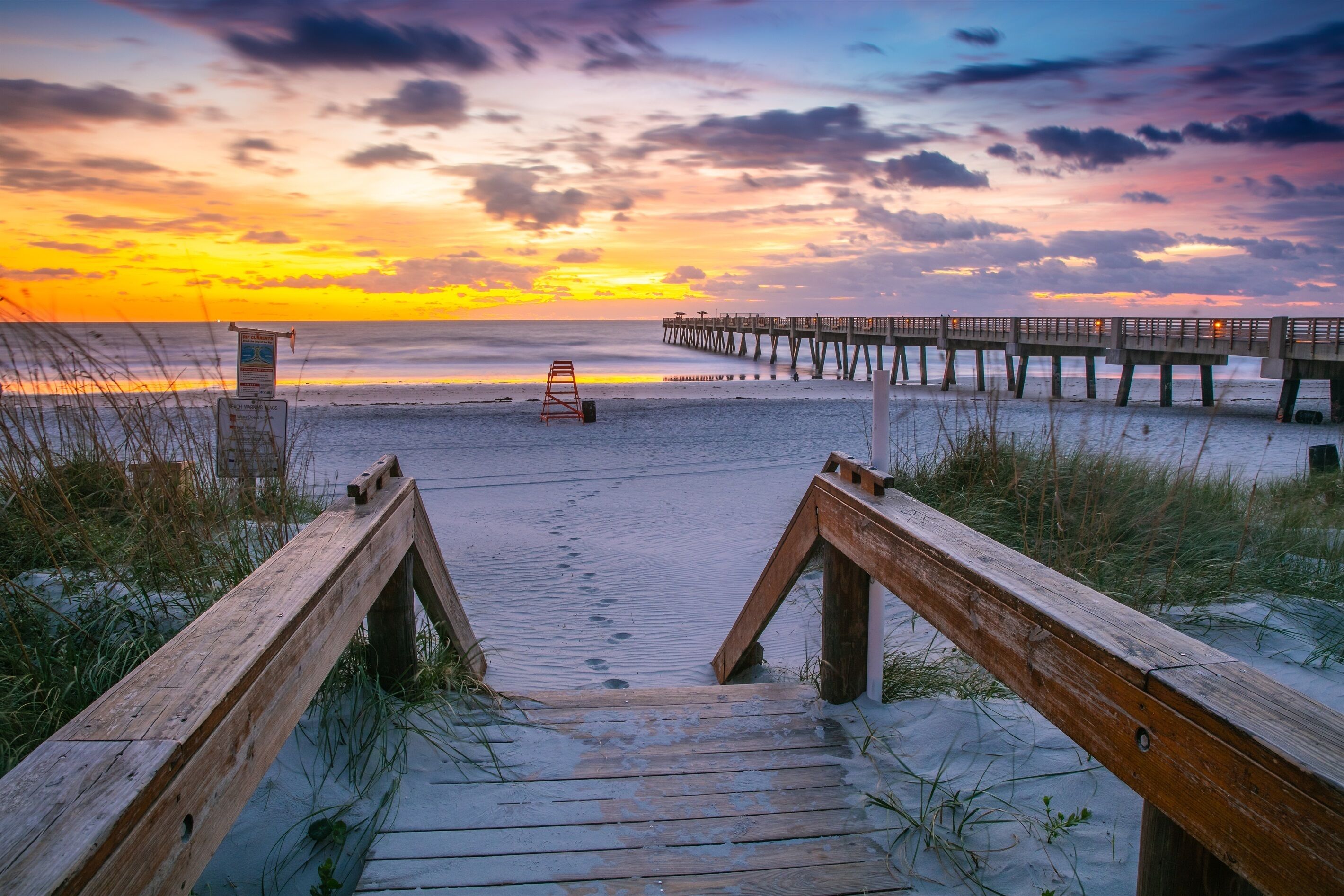 A wooden boardwalk leading to a pier extending into the water, with a lifeguard stand visible on the beach.