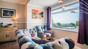 A cozy living room with a wooden cabinet, a cushioned sofa, and a chair with colorful cushions. There is a window with curtains and a view of the outdoors. - Channel View – 3 Bedroom House - Rhossili Bay (Swansea)