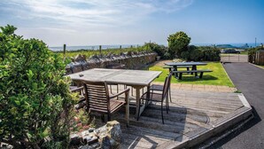 An outdoor patio area with wooden tables and chairs, a stone wall, and a view of the sea. - Channel View – 3 Bedroom House - Rhossili Bay (Swansea)