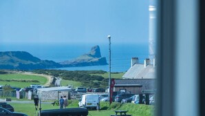 A coastal view with a prominent rock formation, a parking area with cars, and a building with a chimney. - Channel View – 3 Bedroom House - Rhossili Bay (Swansea)
