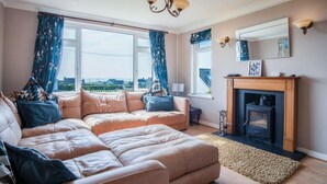 Una sala de estar acogedora con un amplio sofá seccional color beige, una chimenea y vista al mar a través de grandes ventanales.