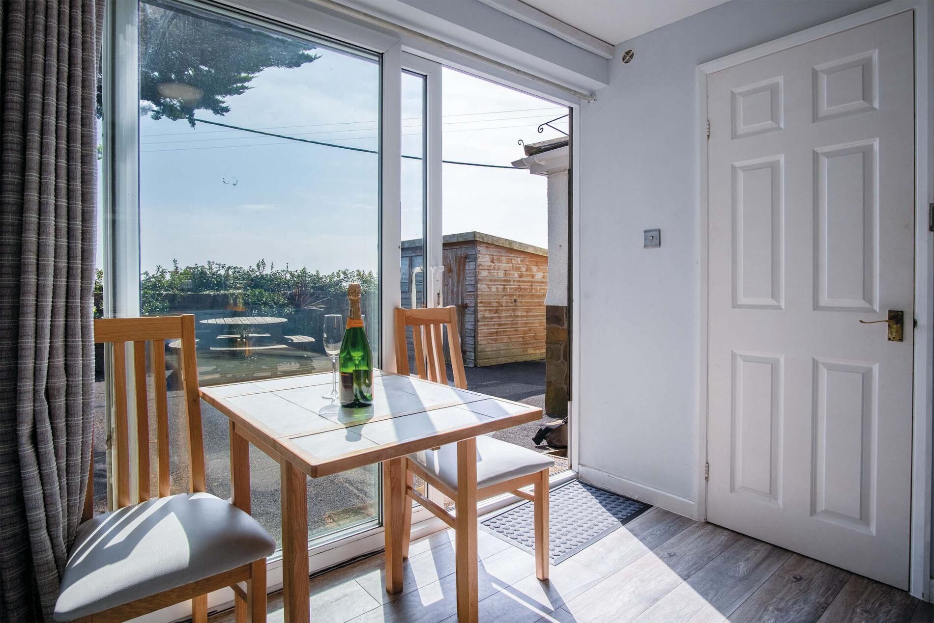 Een kamer met een glazen deur die uitkomt op een terras, een houten tafel met een fles en een glas, en een witte deur.