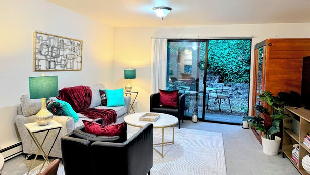 A modern living room with a sofa, armchairs, a coffee table, and a TV stand. There is a sliding glass door leading to a balcony with a view of greenery. - Cedar Heights w sauna/bonfire (Lynnwood)
