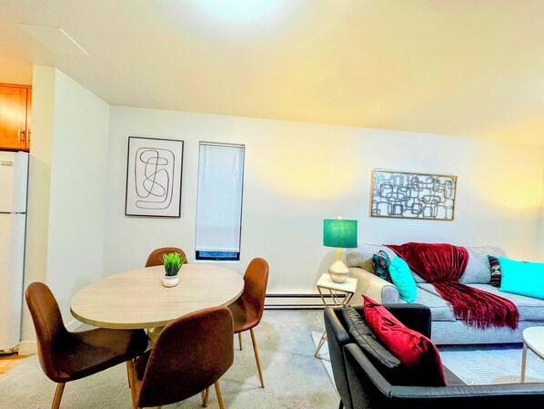 A modern living room with a round dining table, brown chairs, a grey sofa with red and blue cushions, and two wall-mounted abstract art pieces.