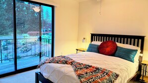 A bedroom with a bed, a ceiling fan, a nightstand, and a view through a sliding glass door to an outdoor area with plants and a fence. - Cedar Heights w sauna/bonfire (Lynnwood)