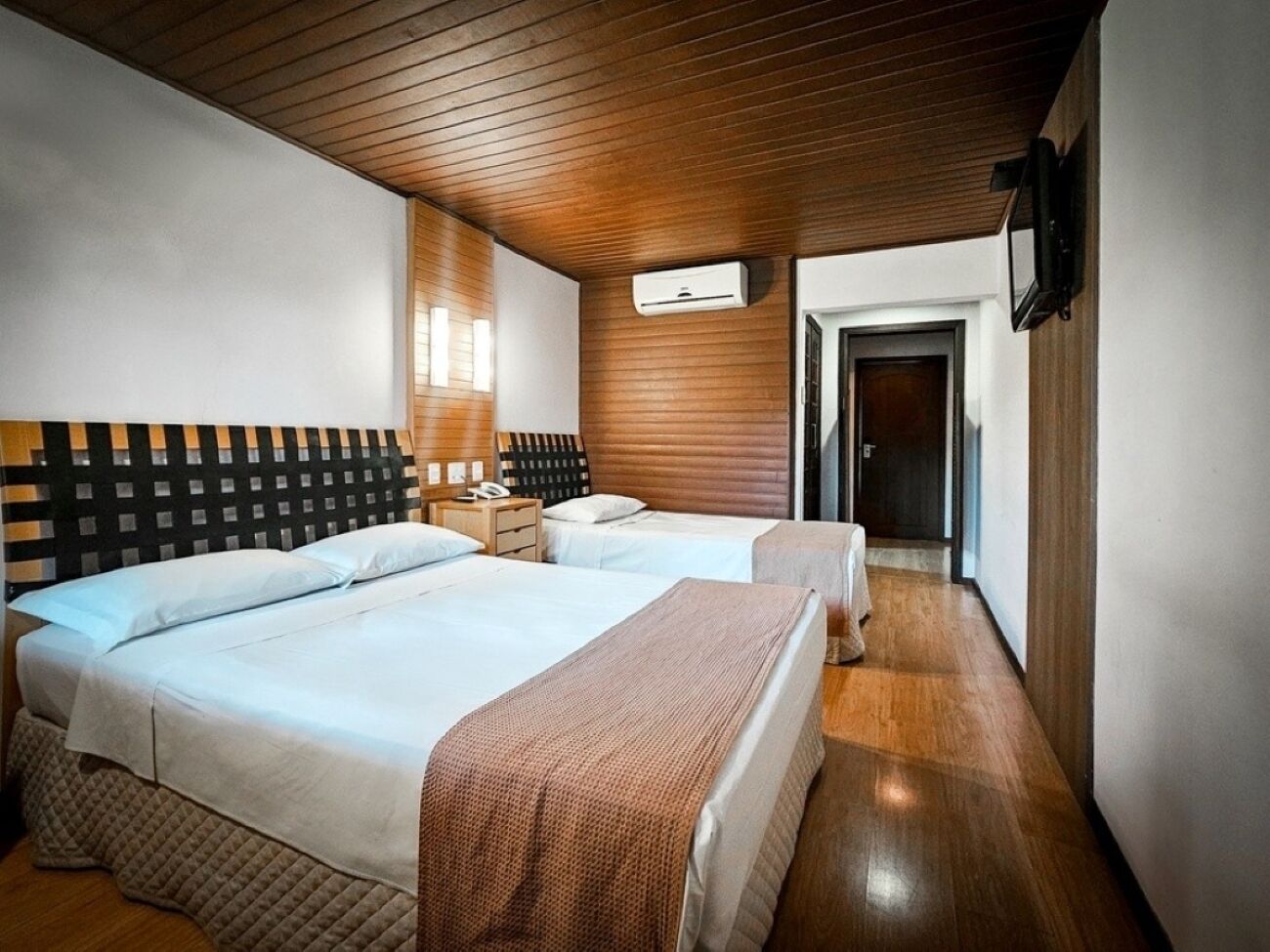 A hotel room with two beds, a wooden ceiling, a wall-mounted air conditioner, a bedside table with a lamp, and a door leading to another room.
