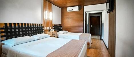 A hotel room with two beds, a wooden ceiling, a wall-mounted air conditioner, a bedside table with a lamp, and a door leading to another room.