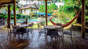 Een overdekt terras met tafels en stoelen, een hangmat en uitzicht op een zwembad en een huis op de achtergrond.