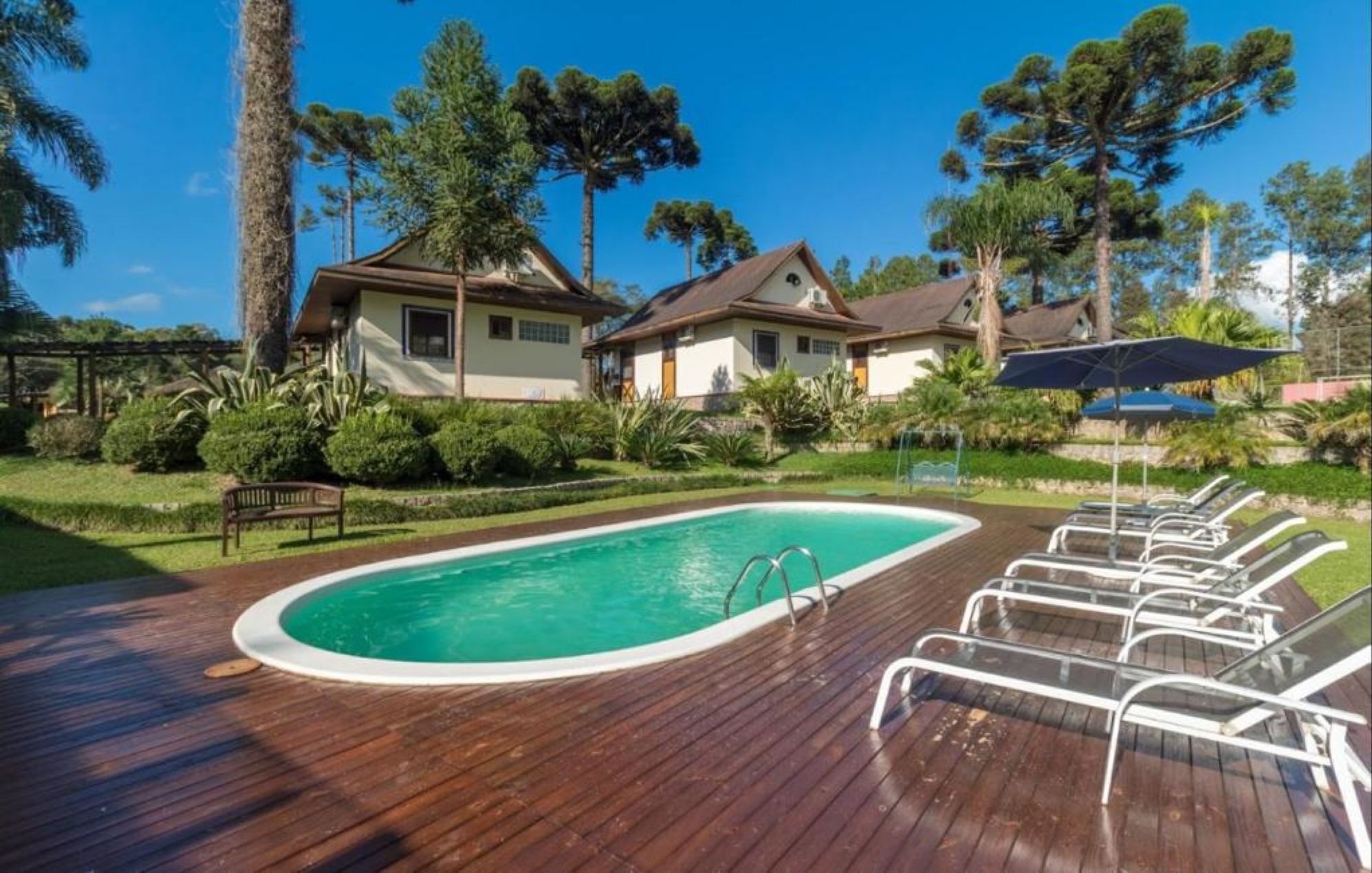 A swimming pool with a wooden deck and lounge chairs, surrounded by greenery and a few houses.
