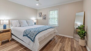 Une chambre à coucher comprenant un lit, des tables de chevet, un ventilateur de plafond, une fenêtre avec des stores, un miroir et une plante en pot.