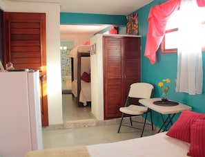 Un dormitorio con cama, una silla, una mesita con un florero, un armario y una puerta que da a otra habitación.