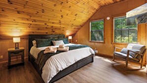 Une chambre confortable avec un plafond en bois, un grand lit doté d’une tête de lit verte, un fauteuil à bascule et une fenêtre donnant sur un paysage verdoyant.