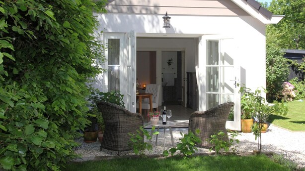 Een huis met een witte gevel, een veranda met rieten meubels en een kleine tuin.