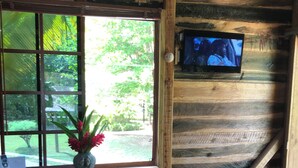A rustic wooden interior with a dining table, two chairs, a vase with flowers, a television on the wall, and a window with a view of greenery.