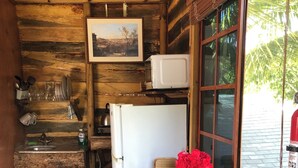 A rustic cabin interior with a wooden bench, a small table, a refrigerator, a microwave, and a window with a view of greenery.