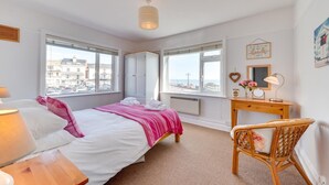 Un dormitorio con cama, mesita de noche, silla, escritorio y ventanas amplias que ofrecen vistas a edificios y al mar.