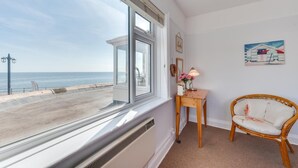 Una habitación con un ventanal grande con vistas a la playa, una silla de mimbre, una pequeña mesa de madera con una lámpara y un cuadro en la pared.