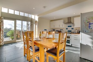 A dining area with a wooden table and chairs, a kitchen with white cabinets, and a large window with curtains. - 3 Bed in Totnes (oc-bcdart) (Broadhempston)