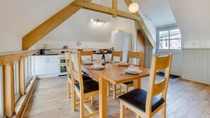 A dining area with a wooden table and chairs, a kitchen in the background, and a sloped ceiling with exposed beams. - 3 Bed in Totnes (oc-bcplym) (Broadhempston)