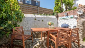 A wooden outdoor table with chairs, a barbecue grill, and a stone wall with a rustic design. - 3 Bed in Brixham (oc-sdbell) (Brixham)