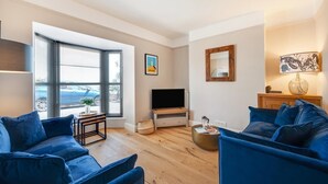 A living room with a TV, blue sofas, a coffee table, and a window with blinds. - 3 Bed in Brixham (oc-sdbell) (Brixham)