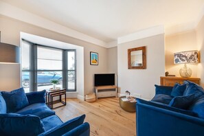 A living room with a TV, blue sofas, a coffee table, and a window with blinds. - 3 Bed in Brixham (oc-sdbell) (Brixham)