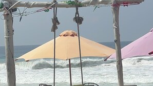 Ένας χώρος με καθίσματα δίπλα στην παραλία, με ξύλινα καθίσματα και τραπέζια, μια κούνια και μια ομπρέλα που λειτουργεί ως στέγαστρο.