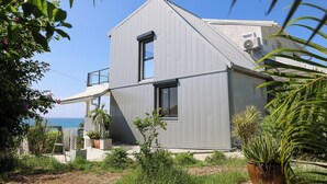 Et moderne hus i to etager med balkon med udsigt over havet, omgivet af frodig vegetation og plantekasser.