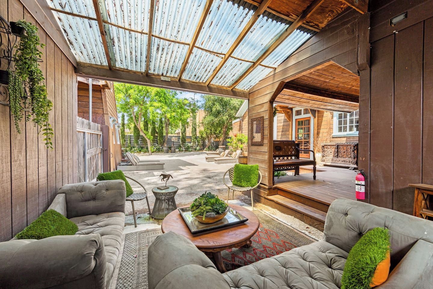 A covered outdoor seating area with cushioned sofas and a coffee table, surrounded by wooden walls and a glass ceiling.