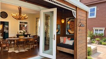 An open-plan living area with a dining table, chairs, a sofa, and a wooden wall with a painting.